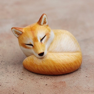 Op de afbeelding: Een klein, opgerold vosje van geverfd klei. De vos is oranje en wit met een gesloten ooguitdrukking.