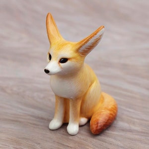 Puede incluir: Una peque&ntilde;a figurita de cer&aacute;mica de un zorro fennec, un zorro del desierto con orejas grandes. La figurita est&aacute; pintada en tonos amarillos y naranjas, con ojos negros y un pecho blanco.