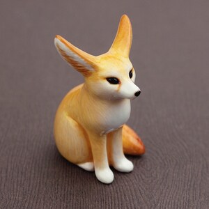 Puede incluir: Una peque&ntilde;a figurita de cer&aacute;mica pintada a mano de un zorro fennec sentado con las orejas levantadas. El zorro es de color marr&oacute;n claro con marcas blancas en la cara y el pecho.
