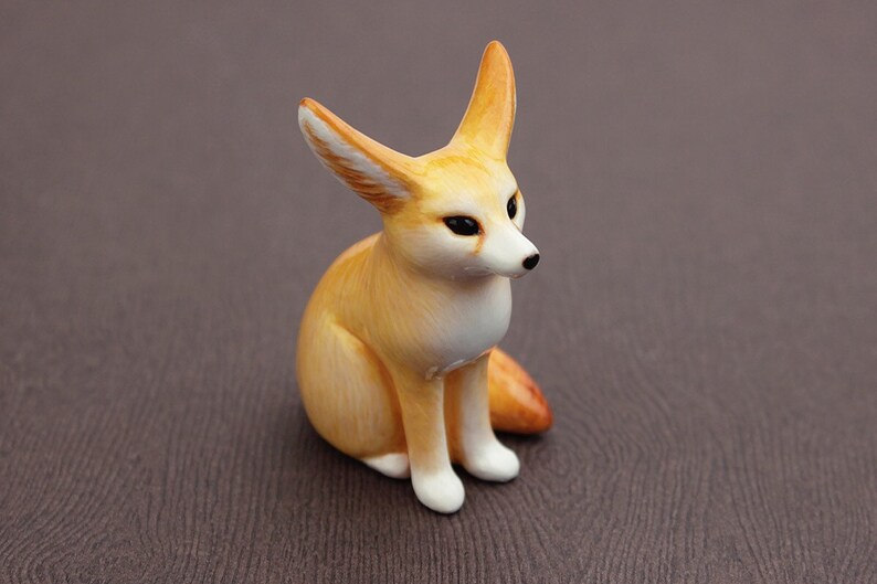 Puede incluir: Una peque&ntilde;a figurita de cer&aacute;mica pintada a mano de un zorro fennec sentado con las orejas levantadas. El zorro es de color marr&oacute;n claro con marcas blancas en la cara y el pecho.
