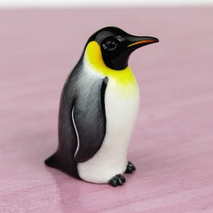 Könnte beinhalten: Eine kleine Keramik-Pinguin-Figur mit schwarz-weißem Gefieder und einer gelben Brust. Der Pinguin steht auf einer rosa Oberfläche.