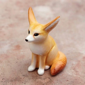 Puede incluir: Una peque&ntilde;a figurita de cer&aacute;mica de un zorro fennec sentado con las orejas levantadas. El zorro es de color marr&oacute;n claro con marcas blancas en la cara y el pecho. La figurita est&aacute; detallada con rasgos realistas, incluida una cola larga y tupida.
