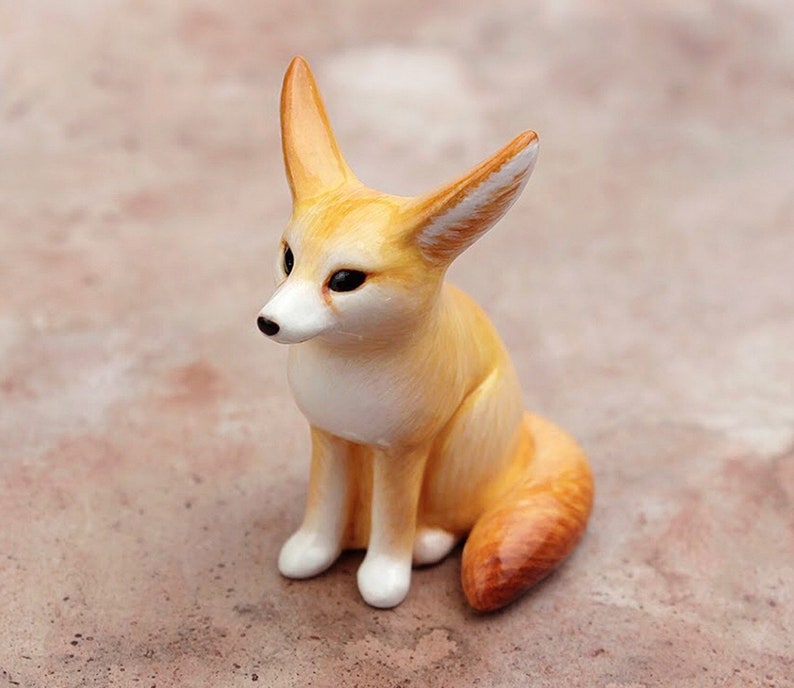 Puede incluir: Una peque&ntilde;a figurita de cer&aacute;mica de un zorro fennec sentado con las orejas levantadas. El zorro es de color marr&oacute;n claro con marcas blancas en la cara y el pecho. La figurita est&aacute; detallada con rasgos realistas, incluida una cola larga y tupida.