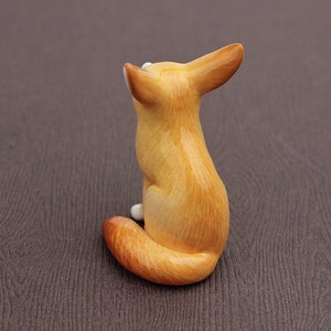 Puede incluir: Una peque&ntilde;a figurita de cer&aacute;mica de un zorro sentado sobre sus patas traseras. El zorro es de color marr&oacute;n claro con el vientre blanco y una cola tupida.