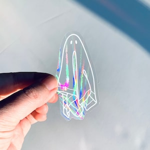 以下が含まれることがあります： 笑顔の顔をした幽霊の白い輪郭が描かれた透明なホログラフィックステッカー。ステッカーは虹色の虹色の仕上げになっています。