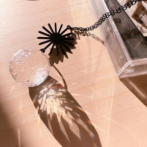 May include: A crystal ball with a black starburst design hanging from a chain. The crystal ball is casting a rainbow-colored shadow on a light brown surface.