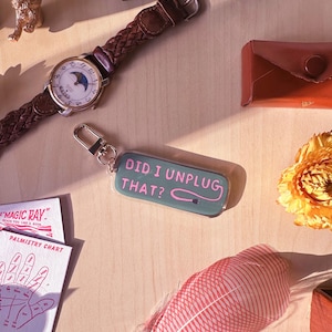 May include: A green and pink keychain with the text "Did I unplug that?"  The keychain is on a wooden surface with a feather, a watch, a small golden lion figurine, and a small brown envelope.
