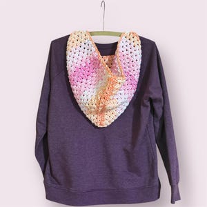 May include: A purple sweatshirt with a crocheted hood. The hood is made of yarn in a gradient of colors, including pink, orange, yellow, and white. The hood is hanging on a green hanger.