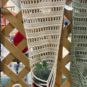 May include: A beige crocheted plant hanger with a small potted plant inside. The hanger is hanging from a wooden lattice.