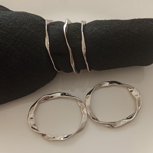 May include: Three silver napkin rings with a wavy, organic design. Two rings are laying flat on a white surface, and one ring is wrapped around a black napkin.