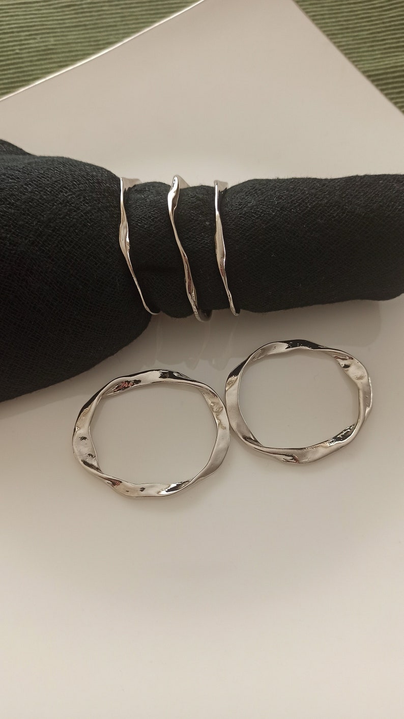 May include: Three silver napkin rings with a wavy, organic design. Two rings are laying flat on a white surface, and one ring is wrapped around a black napkin.