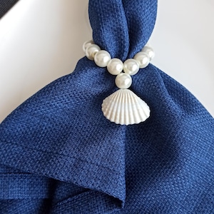 Op de afbeelding: Een marineblauwe linnen servet, gevouwen en vastgehouden door een parelmoeren servetring met een witte schelpenhanger. De opstelling suggereert een nautisch thema.