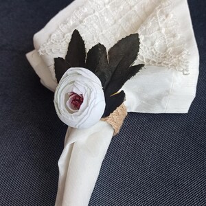 Op de afbeelding: Witte linnen servet met een decoratief bloemaccent. Het accent heeft een witte bloem met een rood hart, donkere bladeren en een met touw omwikkelde ring. De servet heeft een kanten rand.