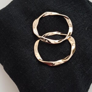 May include: Two gold-toned, twisted hoop earrings. The earrings are on a black fabric background.