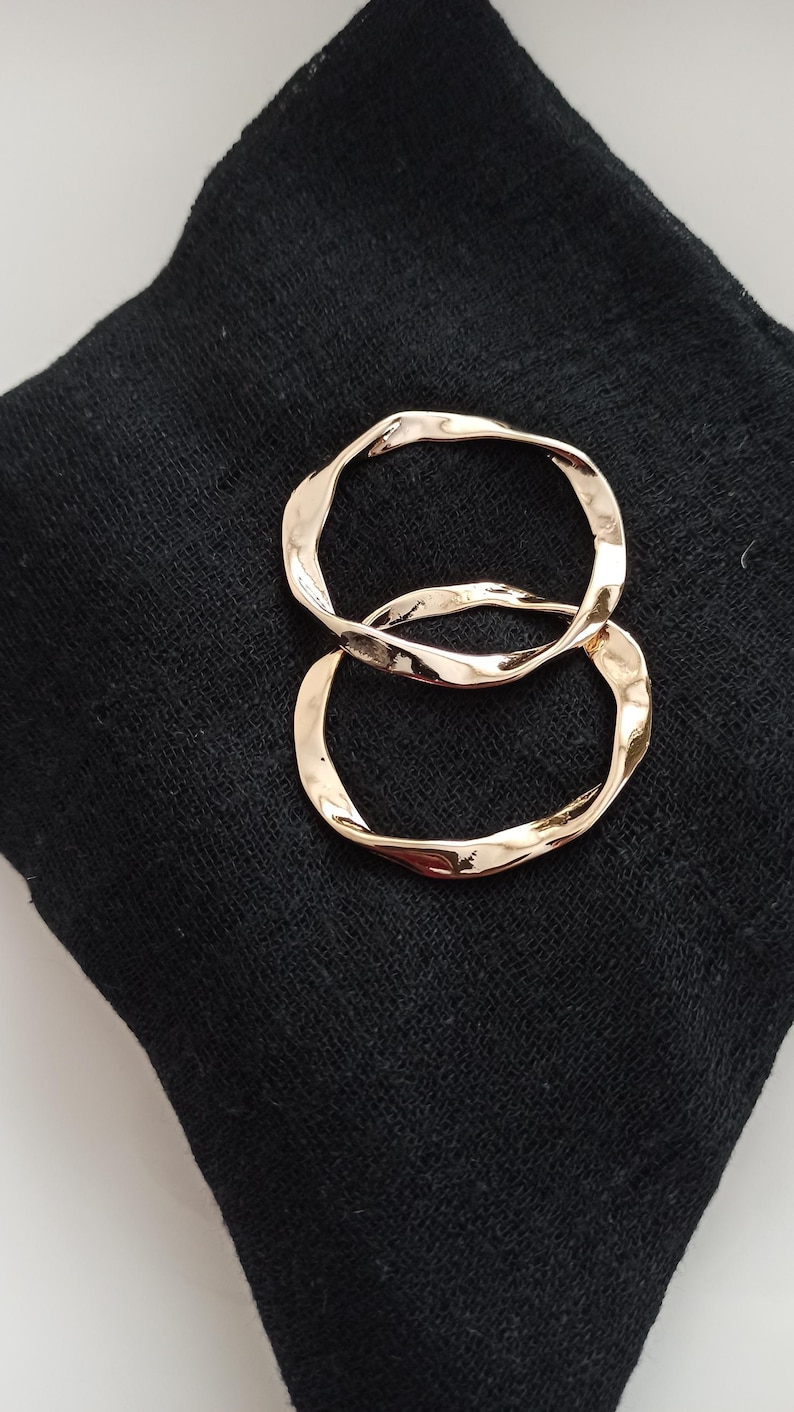 May include: Two gold-toned, twisted hoop earrings. The earrings are on a black fabric background.