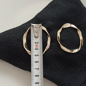 May include: Two gold-toned, twisted, hoop earrings on a black fabric surface. The larger earring is measured with a ruler, showing it is approximately 4.5 inches in diameter.