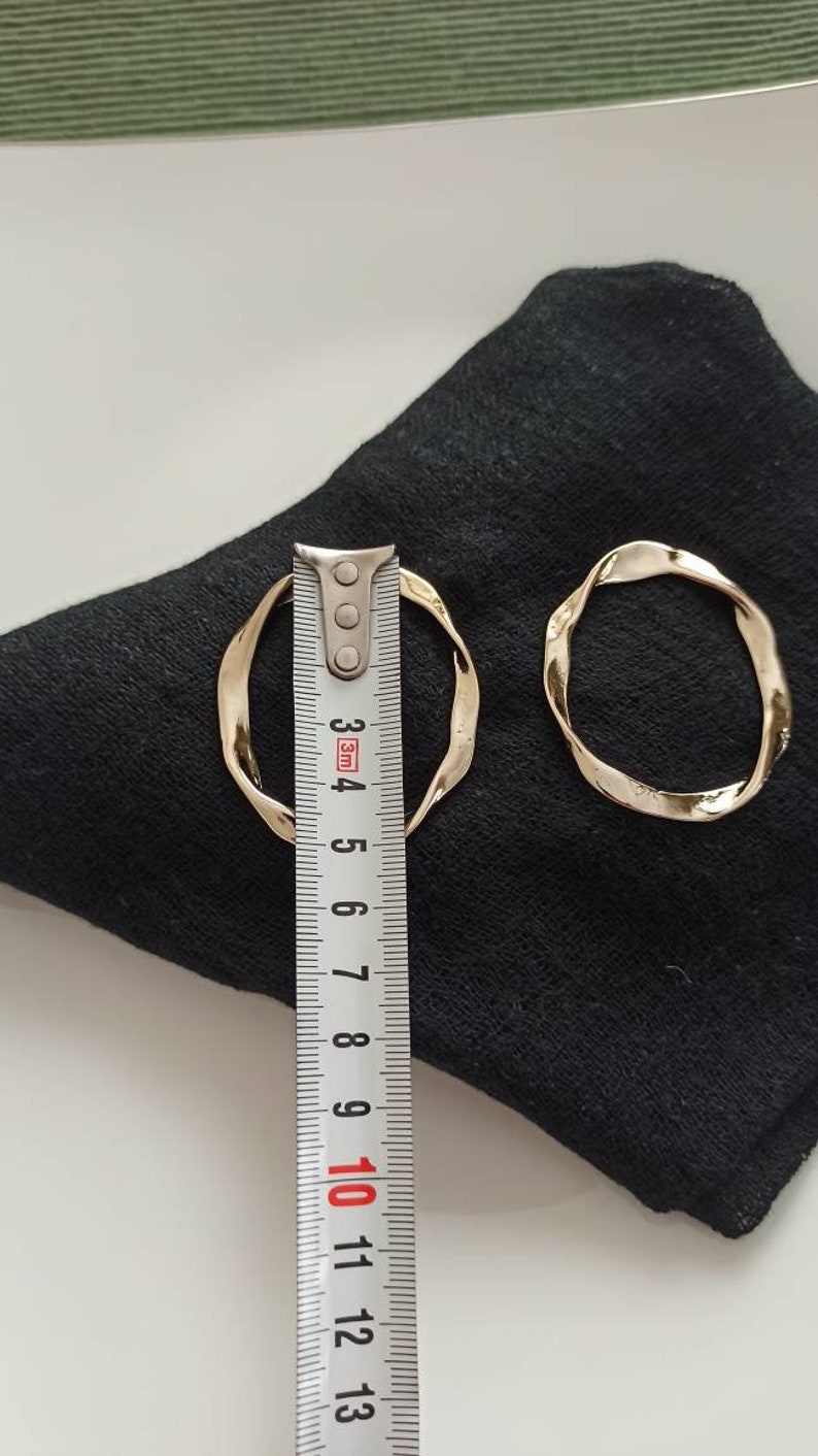 May include: Two gold-toned, twisted, hoop earrings on a black fabric surface. The larger earring is measured with a ruler, showing it is approximately 4.5 inches in diameter.
