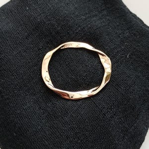 May include: A gold-toned, twisted metal ring, resembling a circle, is displayed on a black fabric background.