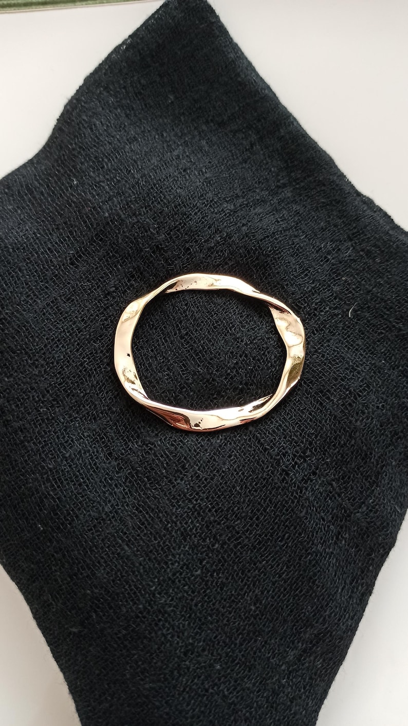 May include: A gold-toned, twisted metal ring, resembling a circle, is displayed on a black fabric background.