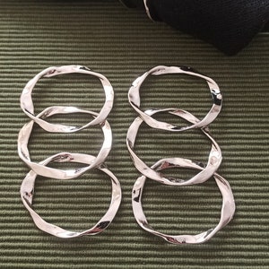 May include: Four silver napkin rings with a wavy, organic design. The rings are stacked on top of each other and are on a green textured surface.