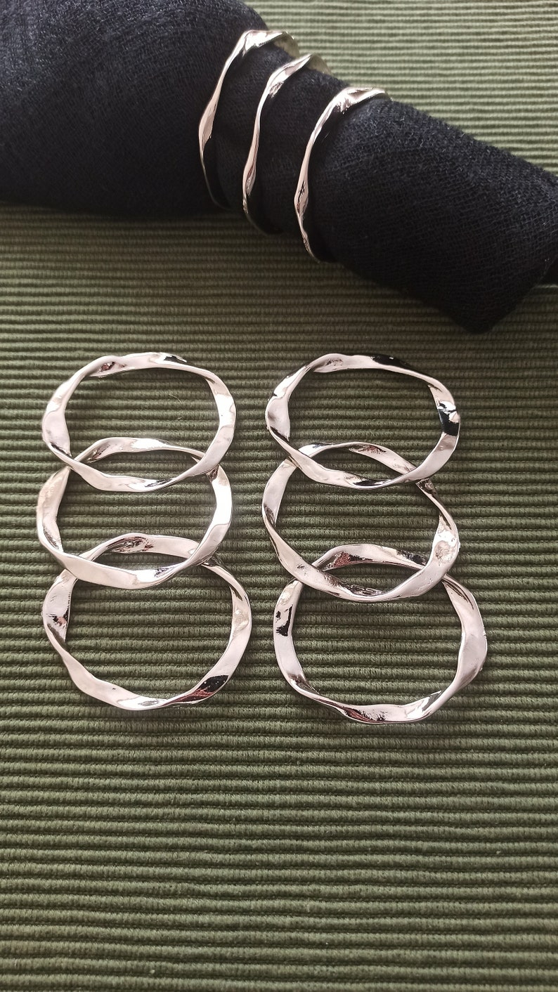 May include: Four silver napkin rings with a wavy, organic design. The rings are stacked on top of each other and are on a green textured surface.