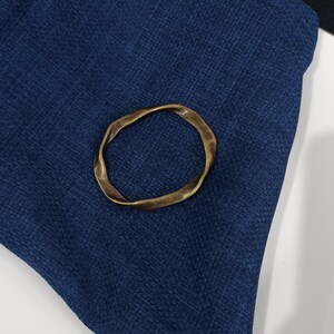 May include: A gold-colored metal ring with an irregular, wavy shape. The ring is lying on a dark blue fabric surface.