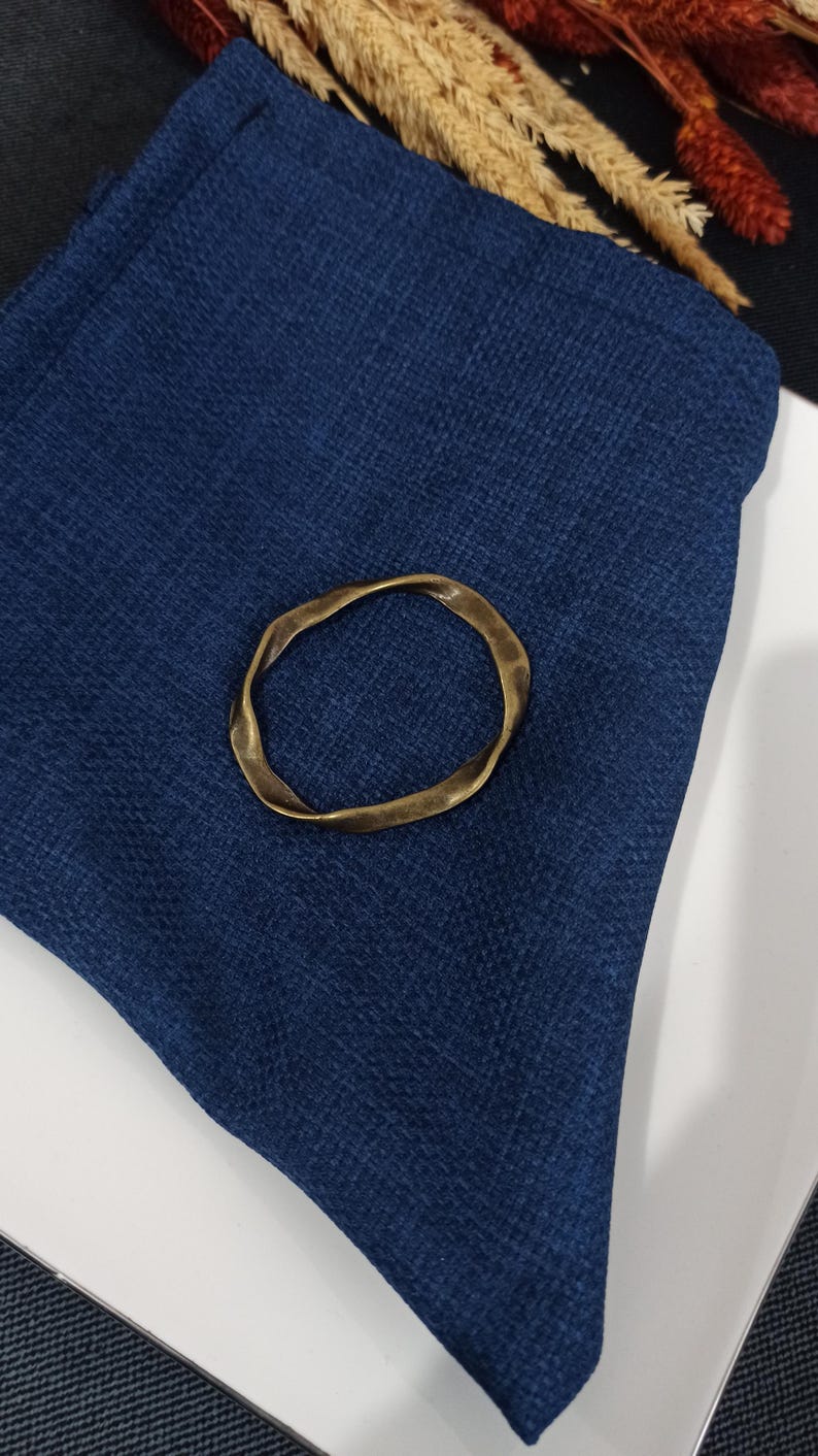 May include: A gold-colored metal ring with an irregular, wavy shape. The ring is lying on a dark blue fabric surface.