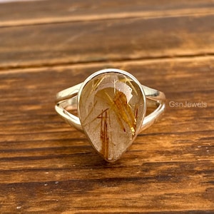 May include: A silver ring featuring a pear-shaped, clear gemstone with golden, needle-like inclusions. The ring has a split shank design and is set against a wooden surface. The text "GsnJewels" is visible on the right side.