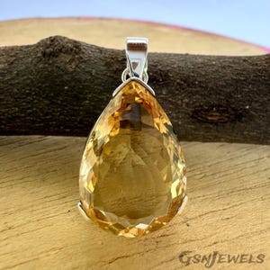 May include: A faceted, teardrop-shaped pendant with a silver bail. The gemstone is a warm, golden-yellow color. The pendant is set against a wooden background. The text "GSNJEWELS" is visible in the lower right corner.