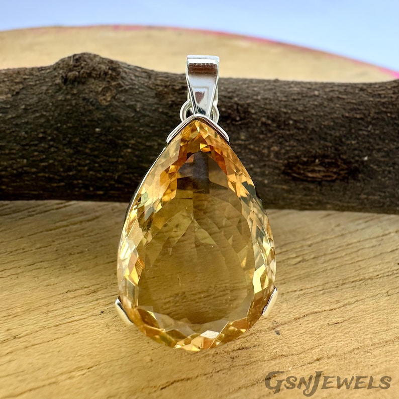 May include: A faceted, teardrop-shaped pendant with a silver bail. The gemstone is a warm, golden-yellow color. The pendant is set against a wooden background. The text "GSNJEWELS" is visible in the lower right corner.
