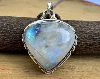 May include: A silver pendant with a large, iridescent moonstone. The stone is a mix of white, blue, and gray, set in a decorative silver frame. The pendant hangs from a thin chain, and the background is a wooden surface.