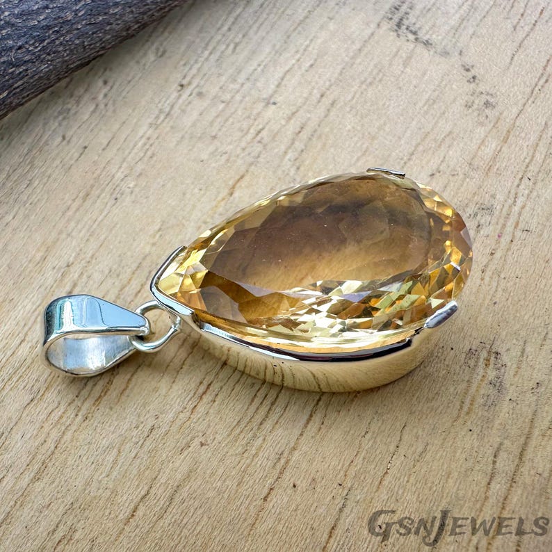 May include: A pear-shaped pendant with a faceted, golden-yellow gemstone set in a silver bezel. The pendant has a silver bail for a chain. The gemstone has a warm, amber hue and is cut to reflect light.