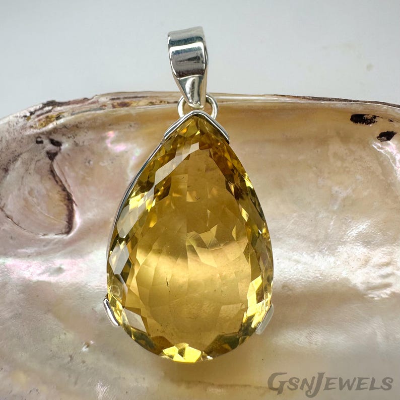 May include: A pear-shaped, faceted citrine pendant with a silver-colored bail. The gemstone is a warm yellow color and is set in a silver-colored metal. The pendant is displayed on a neutral background. The text "GSnJEWELS" is visible.