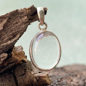 May include: A silver pendant featuring an oval, clear, pale-green gemstone. The gemstone is set in a silver bezel and has a silver loop for a chain. The pendant is shown against a natural wood background.