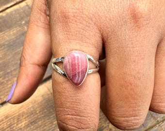 Puede incluir: Un anillo de plata con una piedra preciosa de rodonita rosa en forma de lágrima. La piedra presenta un patrón rayado de tonos rosas claros y oscuros. La banda del anillo tiene un diseño dividido, con dos bandas de plata paralelas.