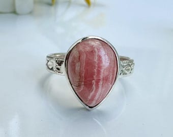 Puede incluir: Un anillo de plata con una gran gema rosa en forma de lágrima con vetas blancas. La gema está engastada en un bisel de plata y la banda tiene un diseño decorativo. El anillo se muestra sobre una superficie blanca.