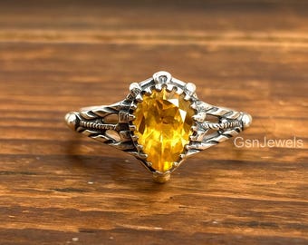 May include: A silver ring with a pear-shaped, bright yellow gemstone. The ring has an ornate, detailed design around the stone and along the band. The text "GsnJewels" is visible on the right side of the image. The ring is set against a wooden background.
