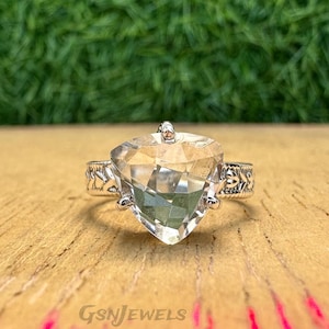 May include: A clear, faceted gemstone ring with a triangular cut. The ring has a silver band with an ornate design. The gemstone is set on a wooden surface. The brand name "GSnJEWELS" is visible at the bottom.