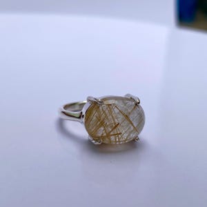 Könnte beinhalten: Ein silberner Ring mit einem klaren, ovalen Edelstein mit goldenen Einschlüssen. Der Edelstein ist in einer Krappenfassung gefasst.