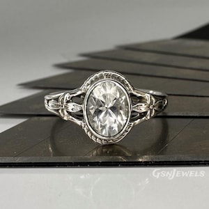 May include: A silver ring with an oval-cut, clear gemstone. The ring has a decorative floral design on the band and around the gemstone setting. The gemstone is the focal point, reflecting light and adding sparkle to the piece.