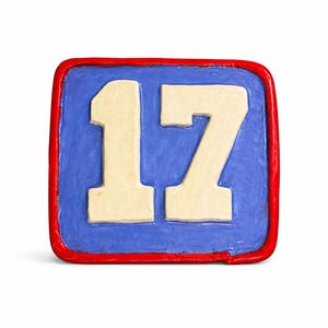 May include: A square, hand-painted wooden block featuring the number 17. The number is cream-colored, set against a blue background, and framed by a red border. The block is on a white background.