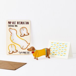 May include: A pop-out sausage dog decoration, a greeting card with a repeating dachshund pattern, and a brown envelope. The pop-out dog is wearing a yellow sweater and red bow tie. The card has blue and yellow dachshunds.