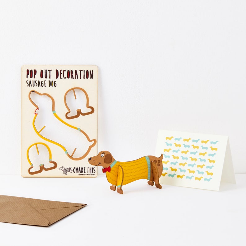 May include: A pop-out sausage dog decoration, a greeting card with a repeating dachshund pattern, and a brown envelope. The pop-out dog is wearing a yellow sweater and red bow tie. The card has blue and yellow dachshunds.