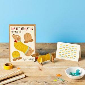 May include: A wooden dachshund pop-out decoration with a yellow sweater and red bow tie, next to a card with the text "Pop Out Decoration Sausage Dog". A matching card with dachshunds in yellow and blue is also present.