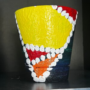 Könnte beinhalten: Handbemalter Blumentopf. Er hat eine strukturierte gelbe Mitte, die von roten und orangen Abschnitten begrenzt und mit weißen Punkten umrandet ist. Der Sockel ist dunkelblau und schwarz.