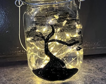 May include: A clear glass mason jar with a silver lid and wire handle, filled with fairy lights. The jar is decorated with a black painted tree design. The lights illuminate the tree and the jar's contents.