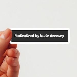 Op de afbeelding: Een rechthoekige sticker met een zwarte achtergrond en witte tekst die luidt: "Geradicaliseerd door basis fatsoen". De sticker wordt tussen vingers tegen een witte achtergrond gehouden.
