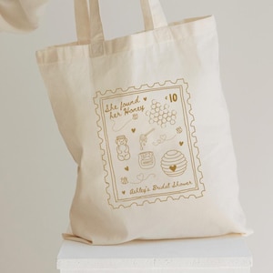 May include: Cream-colored tote bag with a gold-colored stamp design. The stamp reads "She found her Honey" and features honey-themed illustrations. The bag is for Ashley's Bridal Shower.