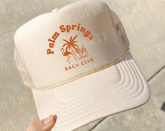 May include: A beige trucker hat featuring a foam front panel and mesh back. The front displays an orange graphic with the text Palm Springs BACH CLUB, a palm tree, a flamingo, and a cocktail glass. A thin braided cord sits above the brim.