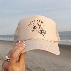 May include: A beige trucker cap with a mesh back and a front panel displaying a dark brown graphic of a cowboy riding a bucking horse. The text KENZIES BACH CLUB is printed in an arc above the illustration, which is accented by two small stars.